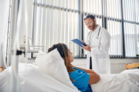 Téléchargez les photos : Médecin indien intelligent parlant à sa patiente afro-américaine couchée dans le lit en salle, soins de santé - en image libre de droit