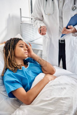 Téléchargez les photos : Malade afro-américaine femme couchée dans le lit à l'hôpital en écoutant son jeune médecin indien - en image libre de droit