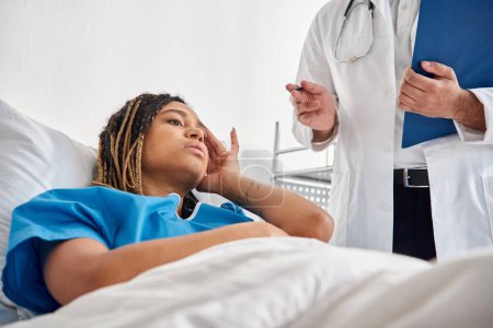Téléchargez les photos : Malade afro-américaine femme couchée dans le lit à l'hôpital en écoutant son jeune médecin indien - en image libre de droit