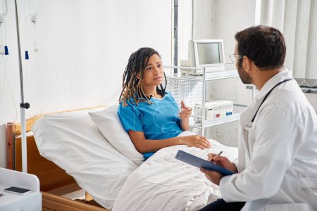 Téléchargez les photos : Attrayant malade afro-américain femme couché dans le lit d'hôpital parler à son jeune médecin indien - en image libre de droit