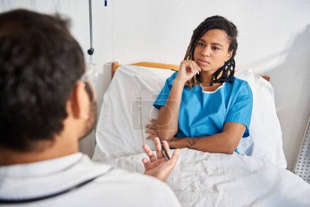Foto de Atractiva mujer afroamericana enferma acostada en la cama de hospital escuchando a su joven médico indio - Imagen libre de derechos