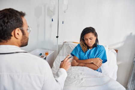 Téléchargez les photos : Attrayant malade afro-américain femme couchée dans le lit d'hôpital à l'écoute de son jeune médecin indien - en image libre de droit