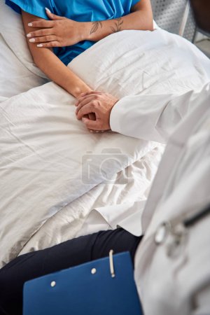 Foto de Joven médico indio sosteniendo la mano del paciente afroamericano mientras yacía en la cama del hospital, recortado - Imagen libre de derechos