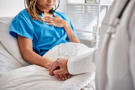 Foto de Vista recortada del joven médico indio sosteniendo su mano de paciente afroamericano acostado en la cama del hospital - Imagen libre de derechos
