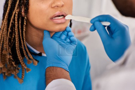 Téléchargez les photos : Vue recadrée du médecin vérifiant la gorge de sa jeune patiente afro-américaine, soins de santé - en image libre de droit