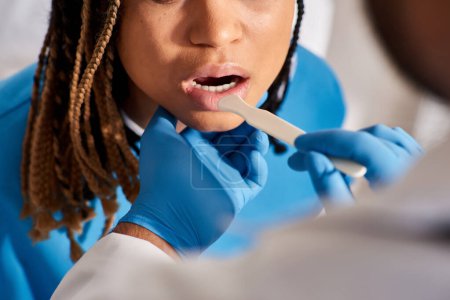 Téléchargez les photos : Vue recadrée du médecin vérifiant la gorge de sa jeune patiente afro-américaine, soins de santé - en image libre de droit