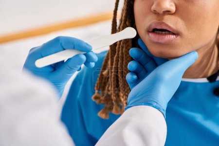 Téléchargez les photos : Vue recadrée de la jeune femme afro-américaine malade en salle d'hôpital ayant médecin contrôle de la gorge - en image libre de droit