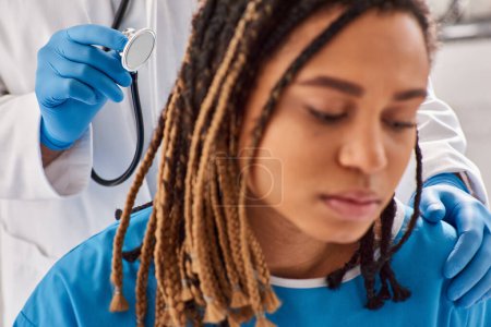 Foto de Enfoque en el médico revisando el aliento de su borrosa paciente afroamericano en la sala de hospital, atención médica - Imagen libre de derechos