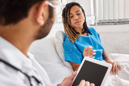 Téléchargez les photos : Afro-américaine patiente couchée dans un lit d'hôpital parlant à son médecin indien prenant des notes sur tablette - en image libre de droit
