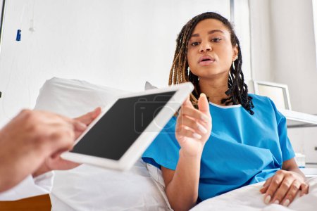 Téléchargez les photos : Focus sur la femme afro-américaine dans le lit d'hôpital en regardant son médecin indien prendre des notes sur tablette - en image libre de droit