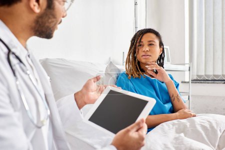 Téléchargez les photos : Focus sur la femme afro-américaine dans le lit d'hôpital en regardant son médecin indien prendre des notes sur tablette - en image libre de droit