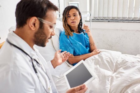 Téléchargez les photos : Focus sur la femme afro-américaine dans le lit d'hôpital en regardant flou médecin indien avec comprimé - en image libre de droit