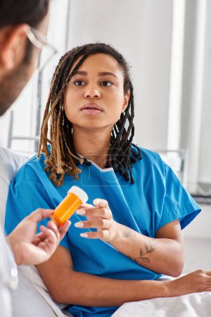 Téléchargez les photos : Se concentrer sur la femme afro-américaine malade dans le lit d'hôpital en prenant la pilule de son médecin indien flou - en image libre de droit
