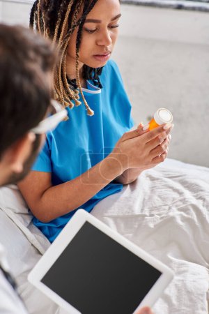 Téléchargez les photos : Se concentrer sur la femme afro-américaine malade assis sur le lit et en regardant ses pilules à côté du médecin indien - en image libre de droit