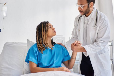 Téléchargez les photos : Médecin indien sérieux réconfortant son patient triste afro-américain dans sa salle d'hôpital, soins de santé - en image libre de droit
