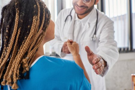 Téléchargez les photos : Vue recadrée du médecin indien joyeux serrant la main de son patient afro-américain joyeux - en image libre de droit