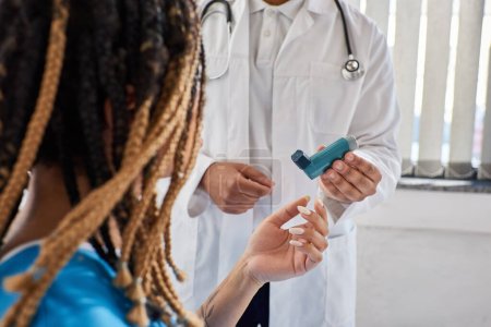 Téléchargez les photos : Jeune médecin indien enseignant à sa patiente afro-américaine malade comment utiliser un inhalateur, recadré - en image libre de droit