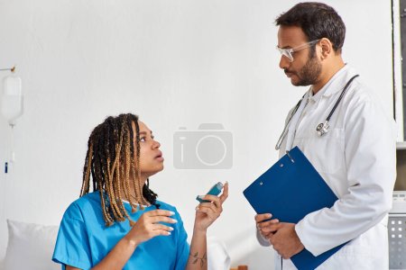 Téléchargez les photos : Jeune médecin indien enseignant à sa patiente afro-américaine malade comment utiliser un inhalateur d'asthme - en image libre de droit
