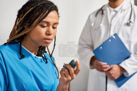 Téléchargez les photos : Focus sur la jeune femme afro-américaine utilisant son inhalateur avec un médecin flou sur fond, soins de santé - en image libre de droit