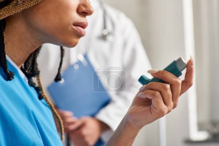 Téléchargez les photos : Vue recadrée de la jeune femme afro-américaine utilisant son inhalateur avec un médecin flou sur toile de fond - en image libre de droit