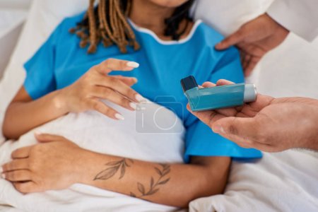 Téléchargez les photos : Vue recadrée de la femme afro-américaine couchée au lit tandis que le médecin indien lui apprend à utiliser un inhalateur - en image libre de droit