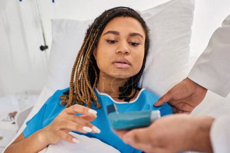 Téléchargez les photos : Se concentrer sur la femme afro-américaine couchée au lit tandis que le médecin indien lui apprend à utiliser un inhalateur - en image libre de droit
