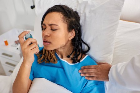 Téléchargez les photos : Jeune femme afro-américaine utilisant son inhalateur d'asthme avec son médecin indien main sur épaule - en image libre de droit