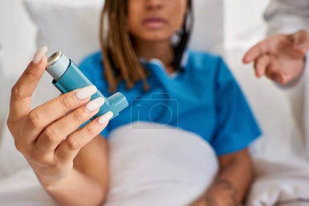 Téléchargez les photos : Se concentrer sur l'inhalateur d'asthme dans les mains d'un patient afro-américain avec un médecin à côté d'elle en salle - en image libre de droit