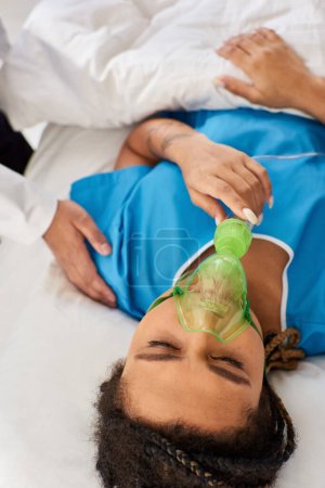 Téléchargez les photos : Vue recadrée du médecin réconfortant femme afro-américaine malade couché dans le lit d'hôpital avec masque à oxygène - en image libre de droit