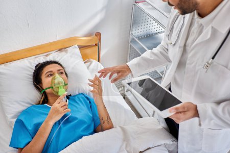 Téléchargez les photos : Malade afro-américaine couchée au lit avec masque à oxygène regardant son médecin indien tenant un comprimé - en image libre de droit