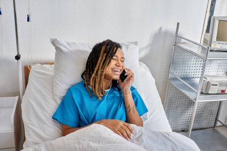 jolly young african american woman talking by phone and smiling in her hospital ward, healthcare