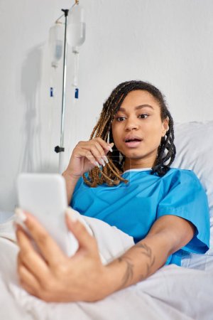 Téléchargez les photos : Séduisante jeune femme afro-américaine ayant un appel vidéo alors qu'elle était allongée dans un lit d'hôpital, soins de santé - en image libre de droit
