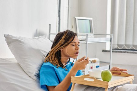 mujer afroamericana joven tomando gachas y manzana para el desayuno en la sala de hospital, atención médica