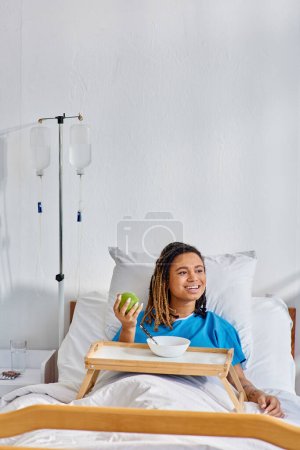 young african american woman having porridge and apple for breakfast in hospital ward, healthcare