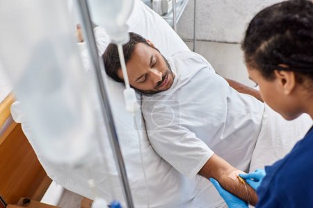 Téléchargez les photos : Jeune infirmière afro-américaine insérant un cathéter dans le bras de sa patiente indienne malade, les soins de santé - en image libre de droit