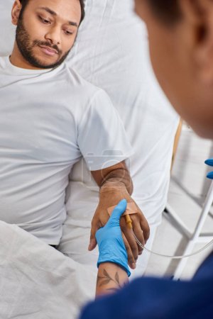 Téléchargez les photos : Vue recadrée de l'infirmière afro-américaine insérant un cathéter dans le bras de sa patiente indienne, les soins de santé - en image libre de droit