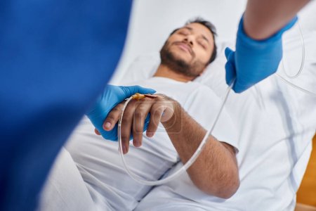 Téléchargez les photos : Focus sur l'infirmière afro-américaine réglage cathéter à côté de l'homme flou couché dans le lit d'hôpital - en image libre de droit