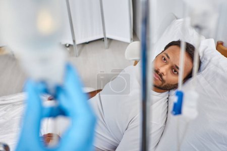 Téléchargez les photos : Se concentrer sur l'homme indien malade couché dans le lit d'hôpital en salle et en regardant la caméra à travers des compteurs de chute - en image libre de droit