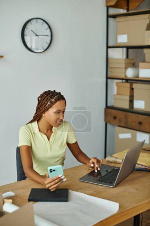 fröhlich attraktive afrikanisch-amerikanische Verkäuferin prüft etwas auf ihrem Laptop und hält ihr Smartphone in der Hand