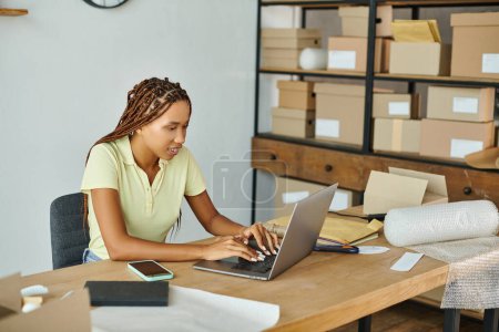joyful african american seller in casual attire working hard on her laptop with parcels on backdrop