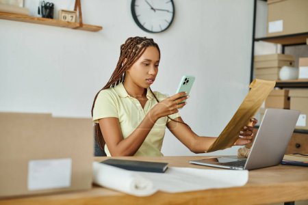 Téléchargez les photos : Séduisante femme afro-américaine tenant téléphone portable et paquet postal dans les mains, concept de livraison - en image libre de droit
