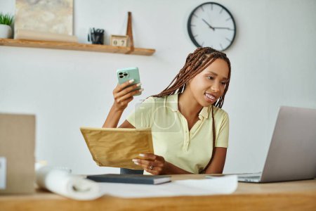 fröhliche junge afrikanisch-amerikanische Händlerin hält Telefon- und Postpaket in der Hand und blickt auf ihren Laptop