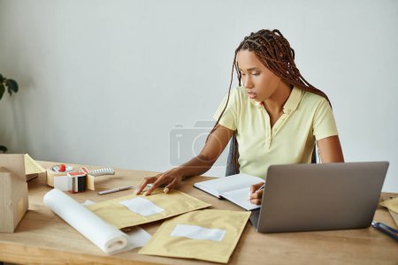 hermosa joven afroamericana vendedora femenina tomando notas mientras trabaja con paquetes de correos