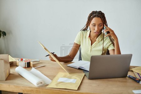 joven africano americano mujer comerciante hablando por teléfono celebración post paquete y mirando portátil