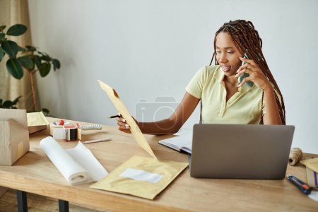 Foto de Alegre atractivo africano americano mujer vendedor hablando por teléfono y mirando post paquete - Imagen libre de derechos