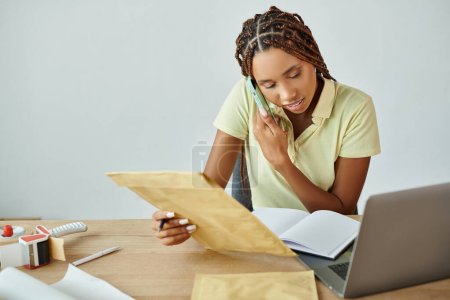 attractive african american seller holding post packet and talking by phone while looking at laptop