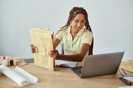 alegre afroamericano comerciante femenino sosteniendo paquetes de correos y mirando a la computadora portátil, entrega