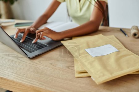 Téléchargez les photos : Vue recadrée de la jeune vendeuse afro-américaine tapant sur son ordinateur portable à côté des paquets de courrier - en image libre de droit