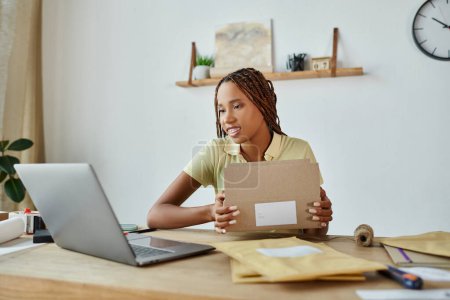 Téléchargez les photos : Belle vendeuse afro-américaine avec bretelles emballage boîte en carton et en regardant son ordinateur portable - en image libre de droit