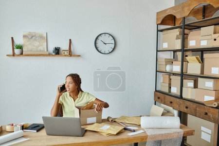 cheerful young african american female seller packing box talking by phone and looking away